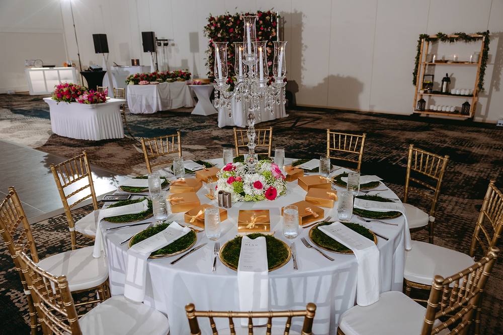 Round reception table with white linen, gold chargers, and a lit crystal candelabra rising above a low pink and white floral centerpiece